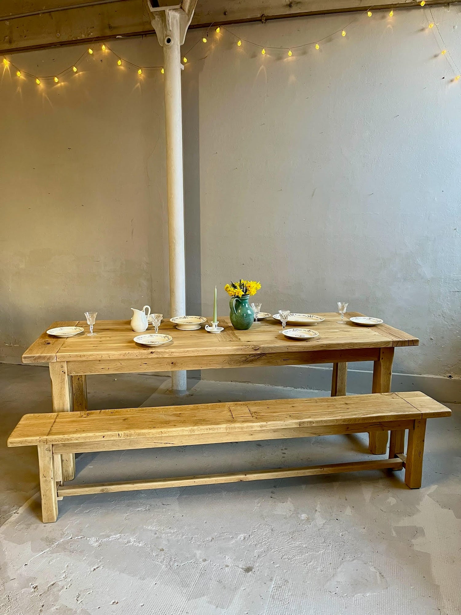 Grande table de ferme en bois brut et son banc assorti, dressés pour un repas avec des assiettes blanches, des verres délicats, un pichet et un vase vert de fleurs jaunes. Une guirlande lumineuse réchauffe le mur gris en arrière-plan.