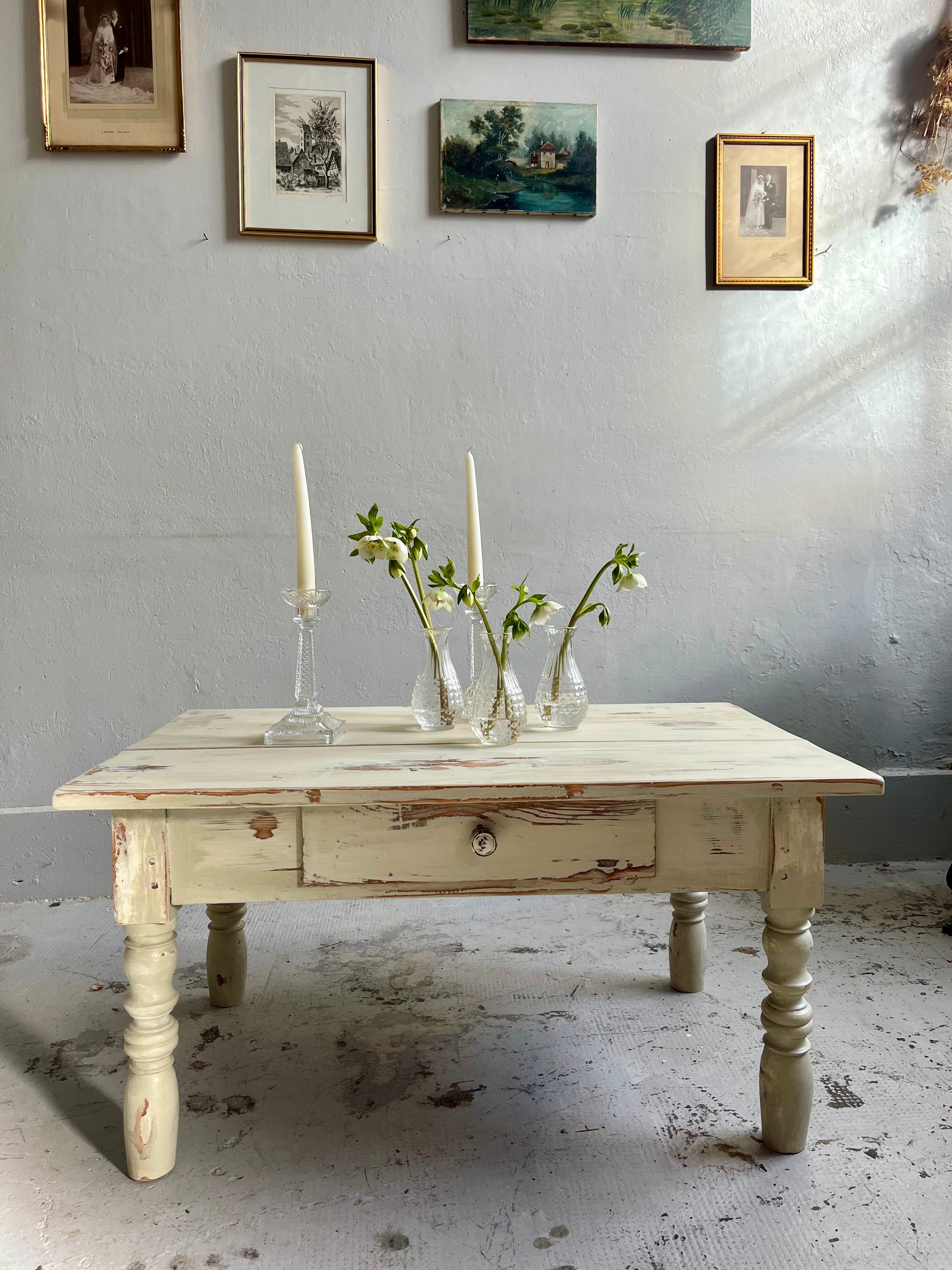 Table basse ancienne en bois à patine crème , dotée d'un tiroir et de pieds tournés, présentée devant un mur gris orné de petits cadres anciens.