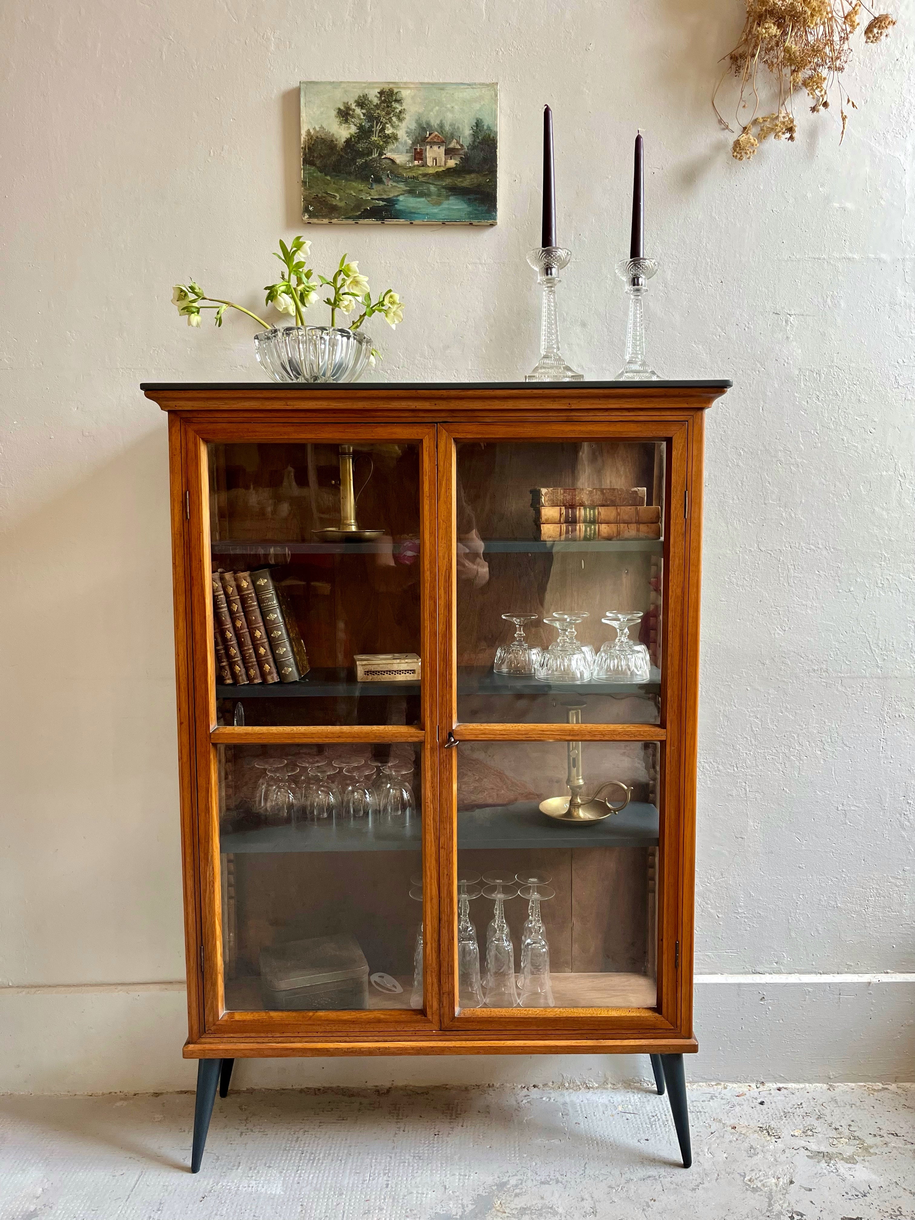 Petite vitrine ancienne en bois avec deux portes vitrées et pieds fuselés noirs, contenant des livres dorés et de la verrerie, surmontée d'un tableau de paysage.