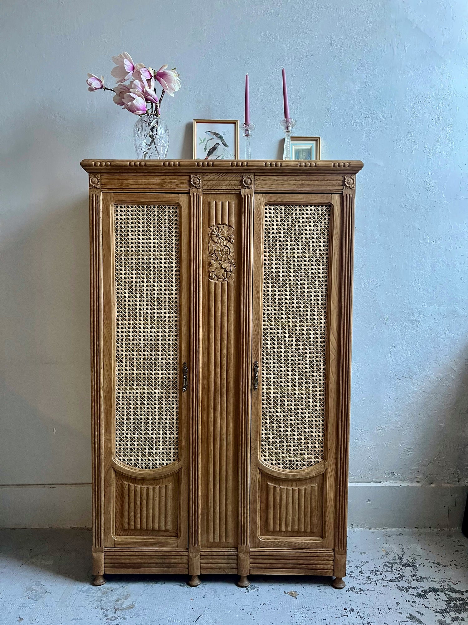 Armoire ancienne en bois sculpté avec portes en cannage rotin, surmontée de fleurs de magnolia dans un vase en verre et de bougies roses.