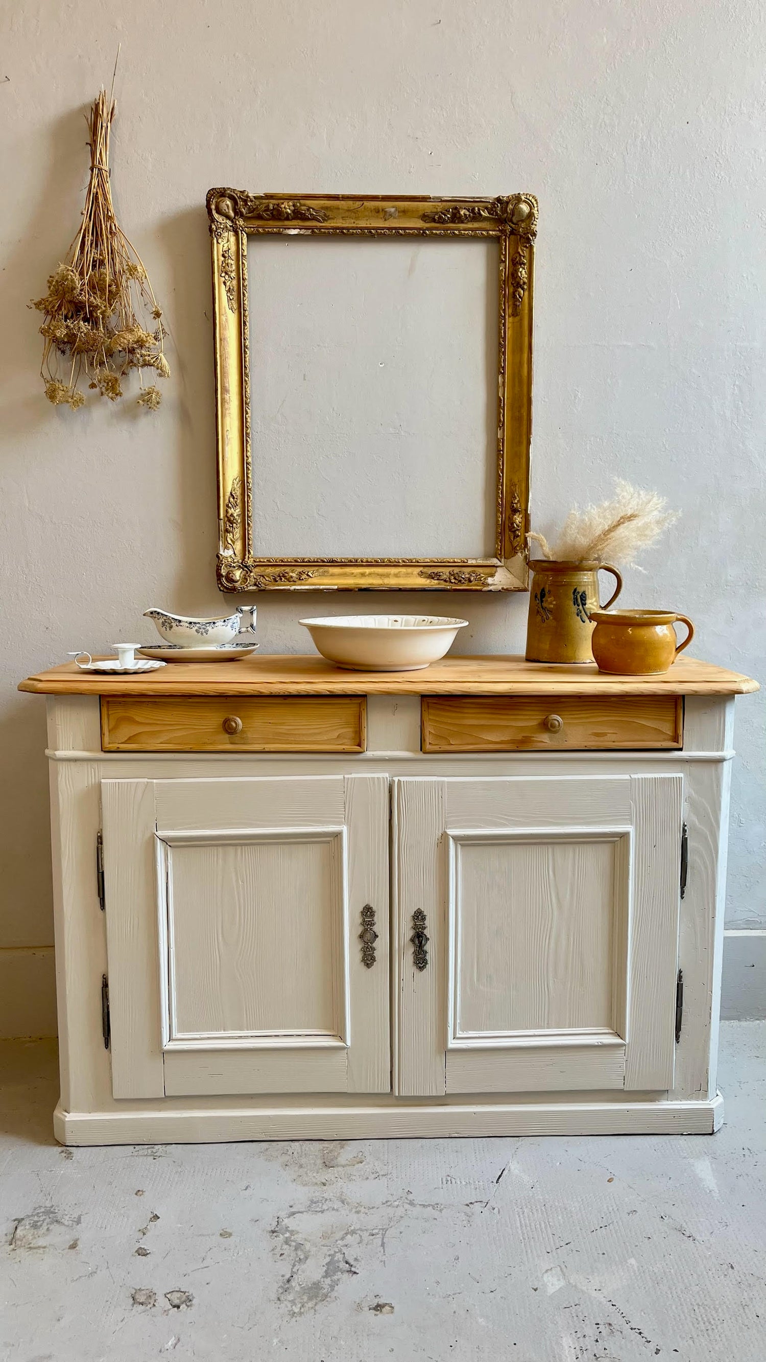 Meuble bas en bois peint en blanc au plateau brut, décoré de vaisselle ancienne et pots en grès. Au mur gris, un grand cadre doré vide et un bouquet séché suspendu complètent cette charmante scène de brocante.