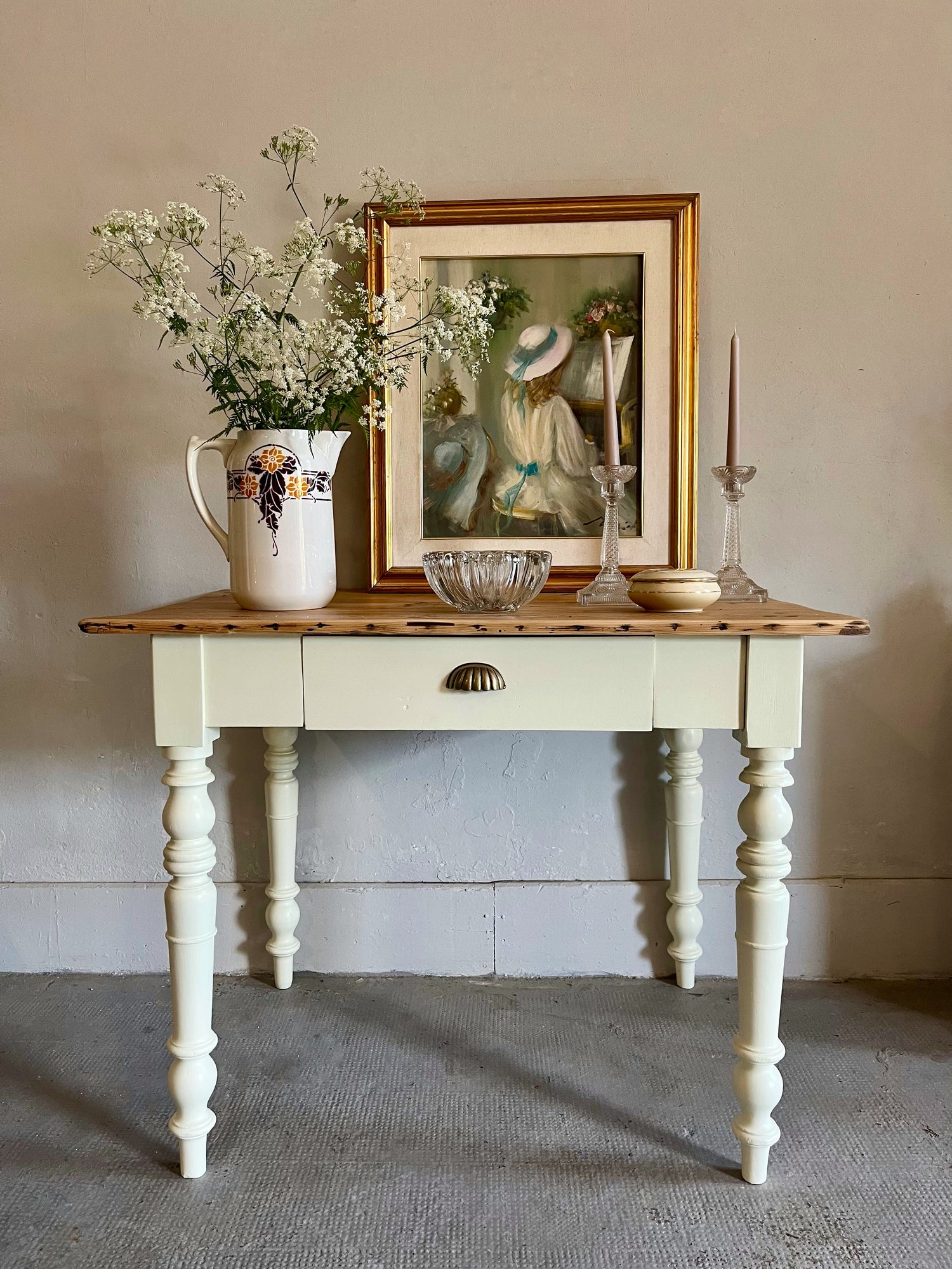 Petite table ou console ancienne en bois peint blanc cassé avec pieds tournés travaillés et un tiroir, décorée d'un grand tableau à cadre doré représentant une femme au chapeau, d'un bouquet de fleurs blanches dans un pichet fleuri et de bougeoirs.