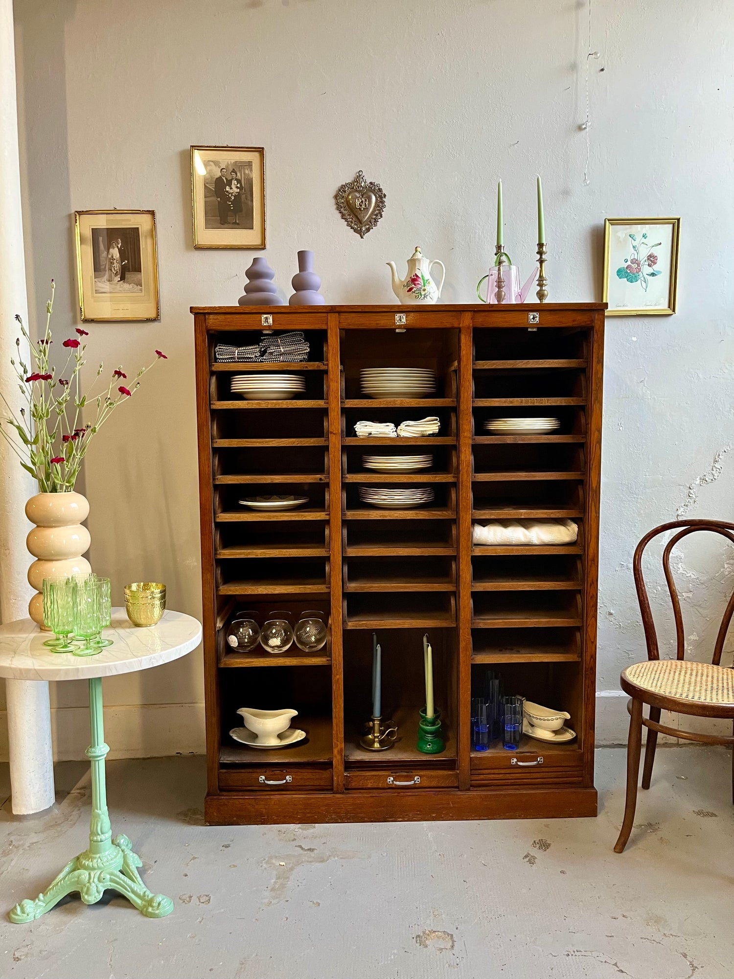 Grand meuble classeur en bois foncé aux rideaux ouverts, révélant des étagères garnies de vaisselle ancienne, verres et bougies. À gauche, un guéridon au piètement vert porte un vase fleuri ; à droite, une chaise en cannage. Des cadres anciens décorent le mur gris.