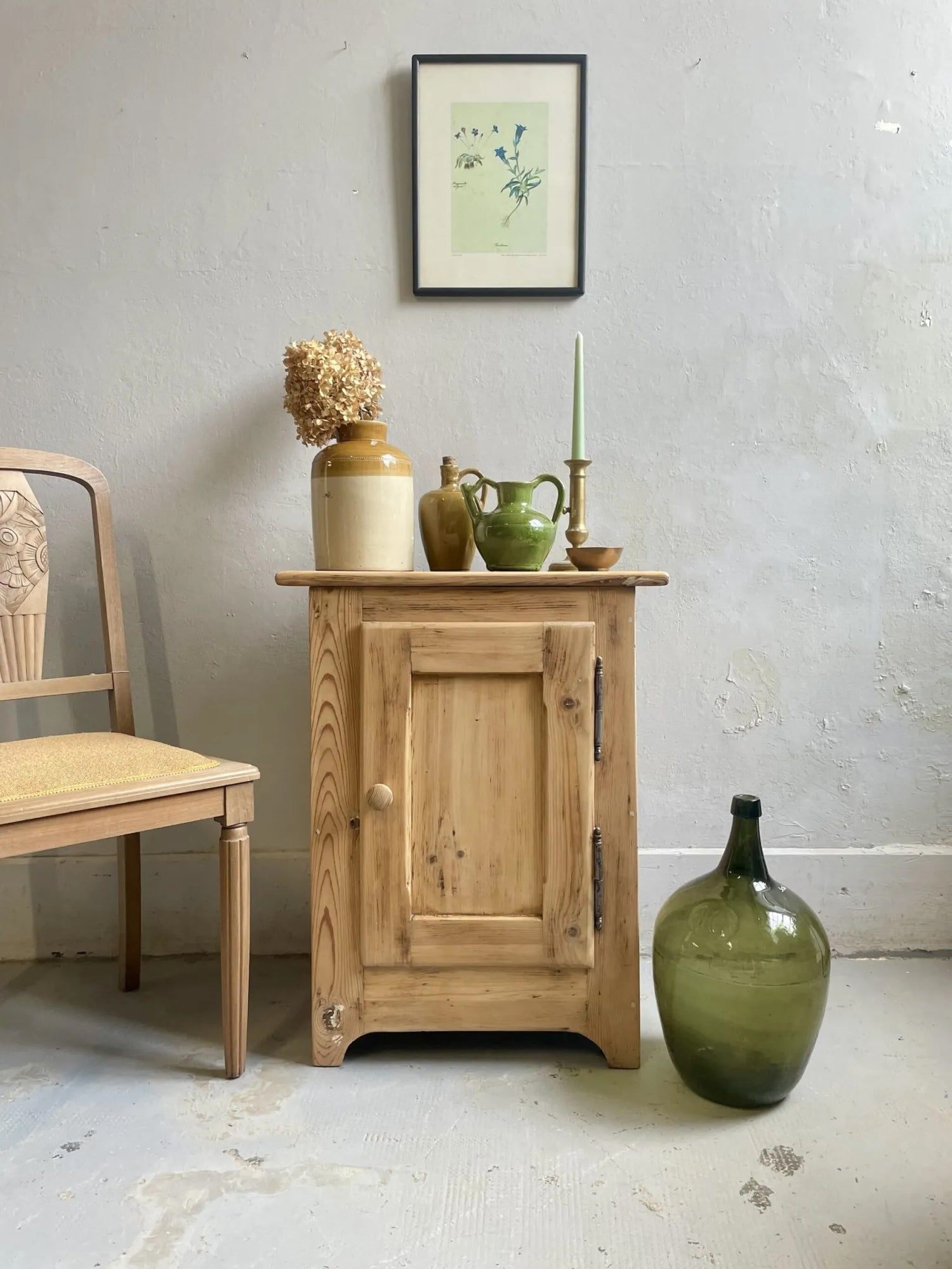 Petit meuble ancien en bois décapé avec porte et ferrures d’origine, mis en scène avec des objets de décoration vintage, une chaise ancienne et une dame-jeanne verte dans un intérieur lumineux.
