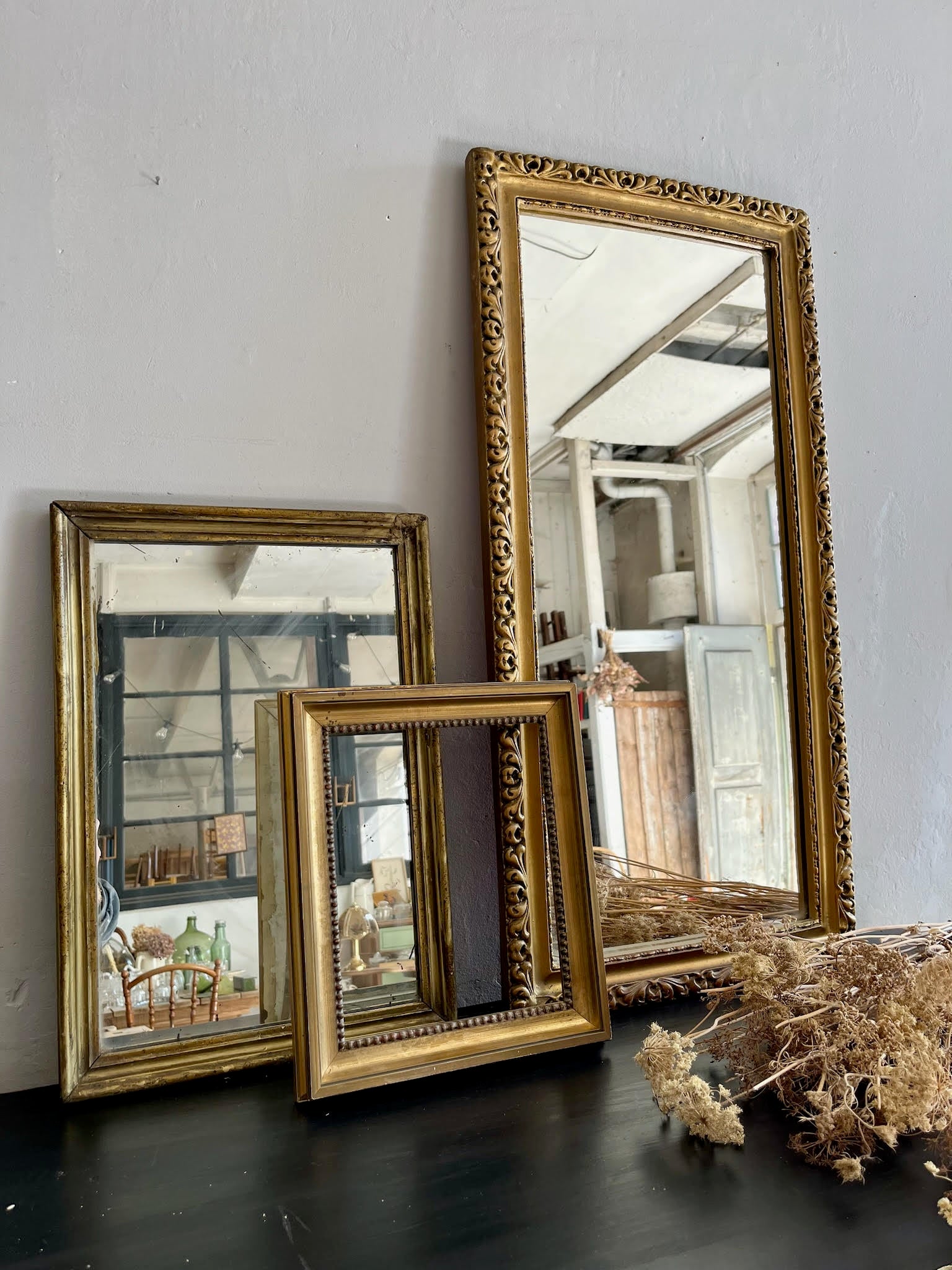 Trois miroirs anciens aux cadres dorés sculptés de différentes tailles, posés sur un meuble noir contre un mur gris. À droite, un bouquet de fleurs séchées apporte une touche naturelle et rétro à cette composition de brocante.