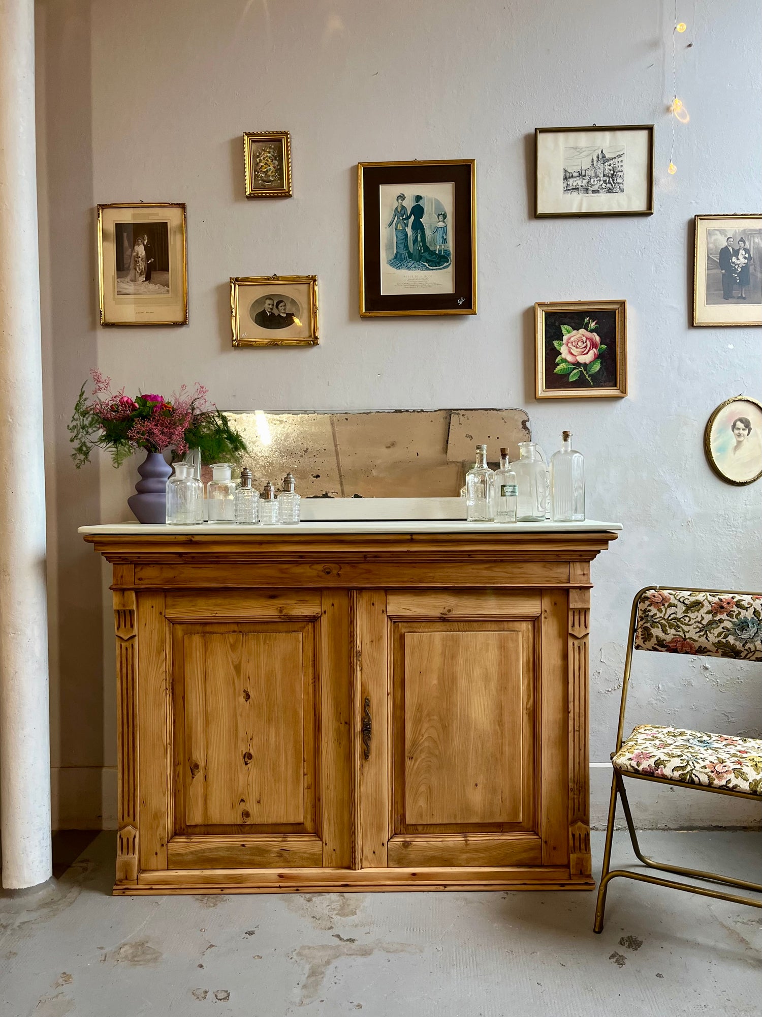 Grand meuble bas en bois brut au plateau de marbre blanc, surmonté d'un miroir ancien piqueté, de flacons en verre transparent et d'un vase garni de fleurs. À droite se trouve une chaise pliante fleurie rétro. Une composition de plusieurs petits cadres anciens orne le mur beige en arrière-plan.