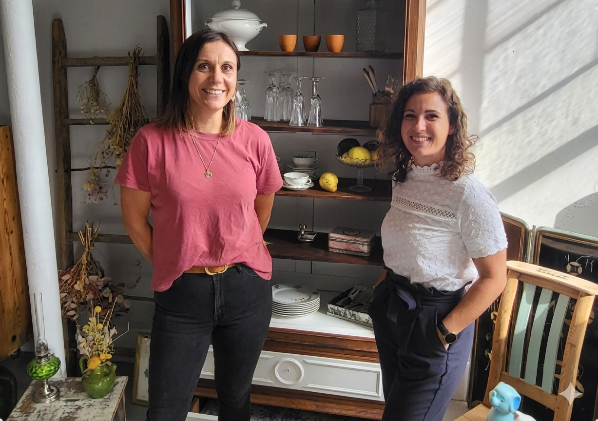 Portrait de deux femmes souriantes posant dans un atelier de décoration intérieure, entourées de mobilier ancien et d'objets artisanaux.