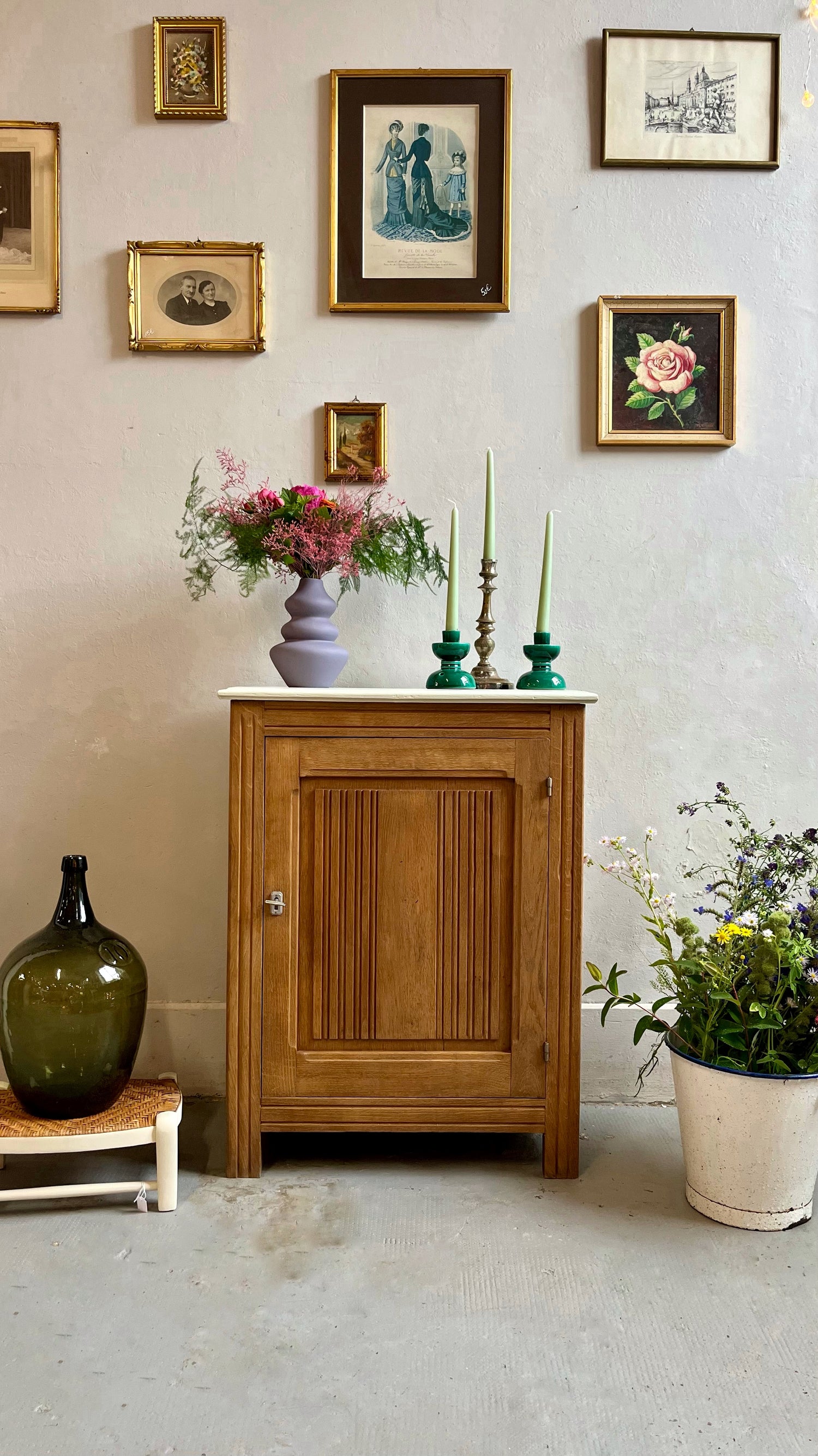 Petit meuble en bois au plateau blanc, surmonté d'un vase de fleurs violettes et de chandeliers verts. À gauche, une dame-jeanne foncée repose sur un petit banc ; à droite, un grand seau de fleurs champêtres. Une composition de cadres anciens orne le mur beige en arrière-plan.