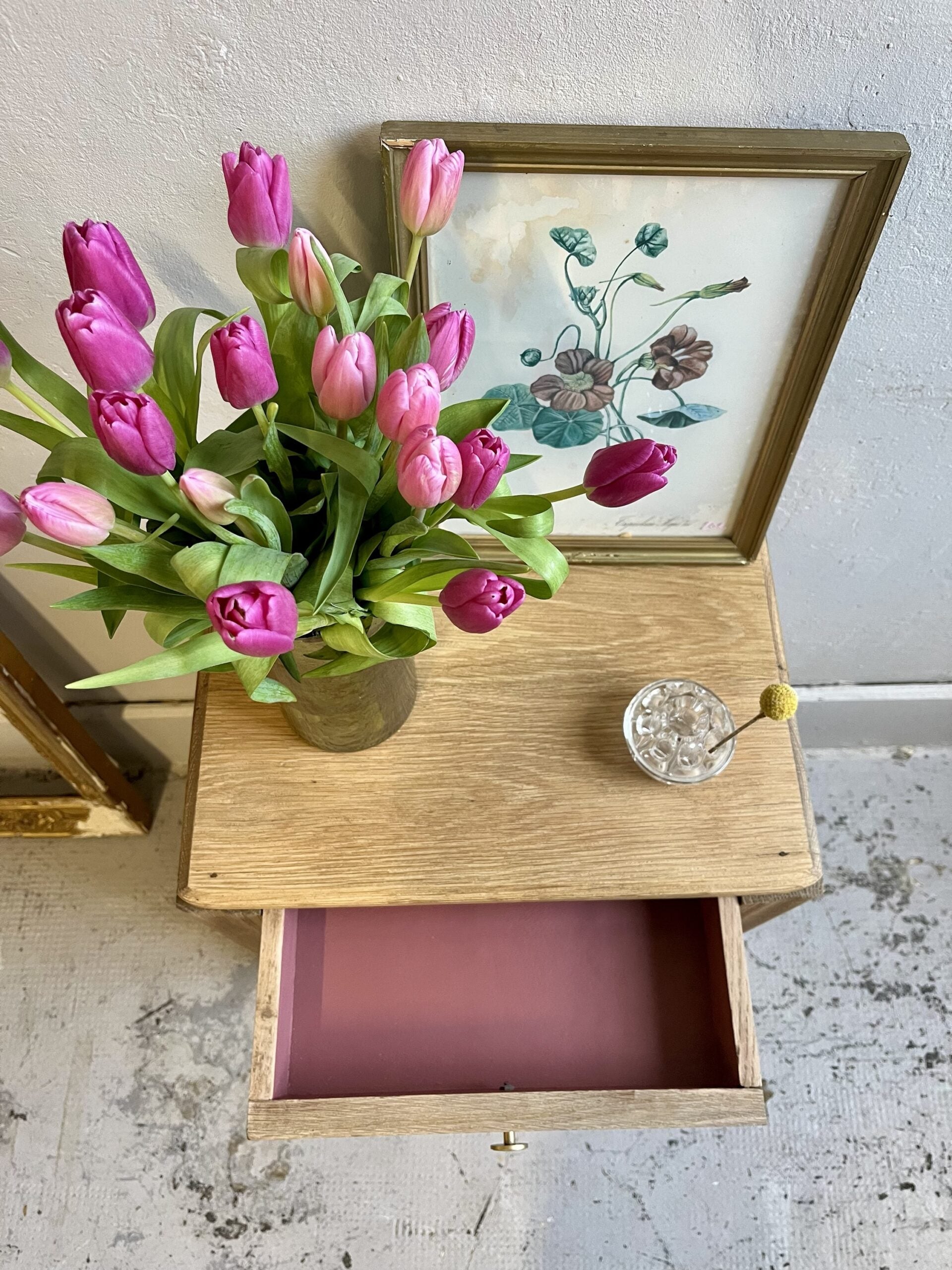 Vue de dessus d'un plateau de chevet en bois clair avec tiroir ouvert révélant un fond rose, décoré d'un bouquet de tulipes violettes et d'un cadre ancien.