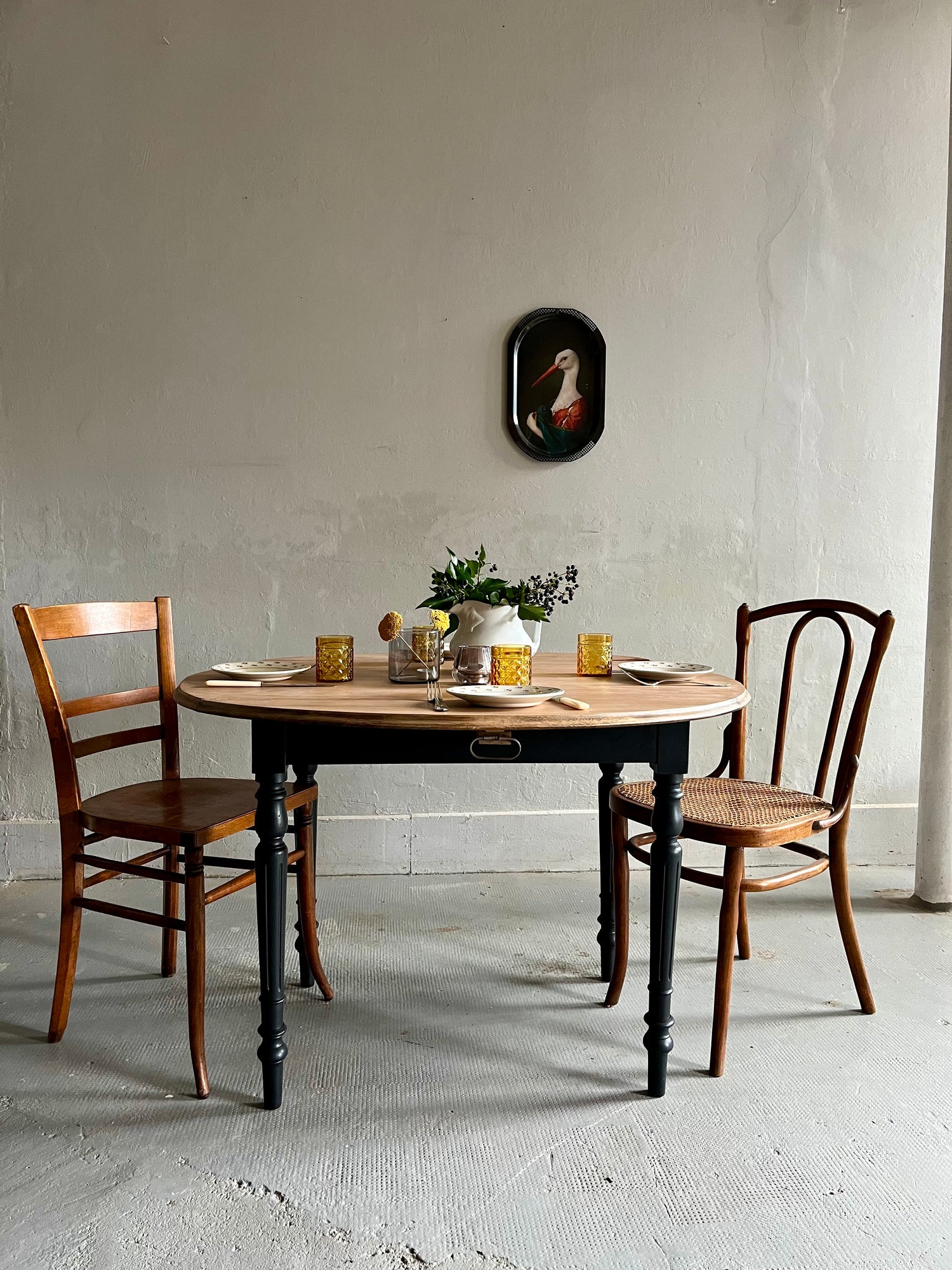 Table ronde ancienne dotée d'un piétement noir et d'un plateau en bois clair, entourée de deux chaises dépareillées en bois. Dressée de façon épurée, elle se détache devant un grand mur gris clair agrémenté d'un petit cadre noir suspendu.